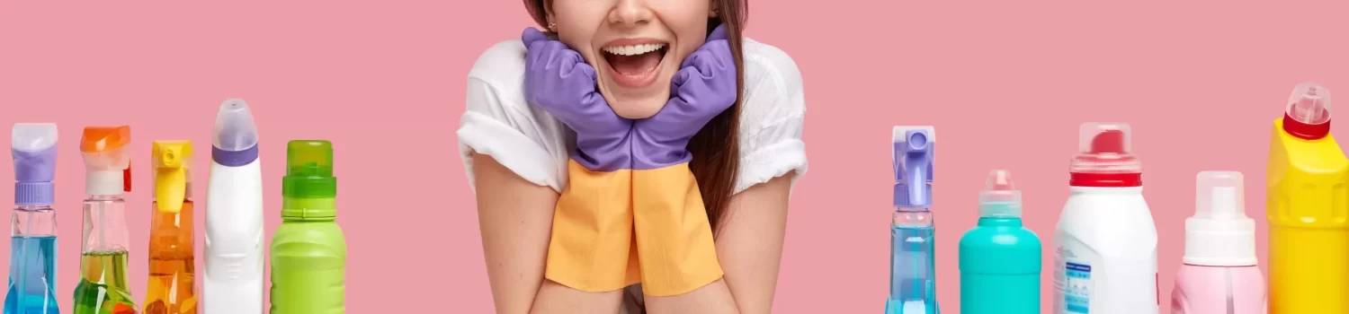 Glad female janitor keeps hands under chin, looks happily away, wears headband and casual t shirt, uses detergents and sponges for cleaning, isolated over pink background. Household concept.